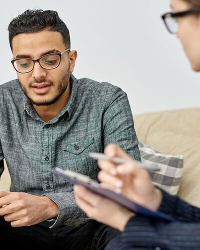 Junger Mann mit Brille sitzt auf einer Couch, neben ihm auf einem Stuhl sitzt eine Frau, die sich Notizen macht