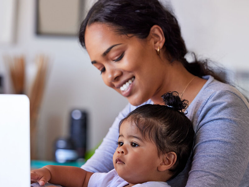 Junge Mutter arbeitet mit ihrem Baby auf dem Schoß am Laptop.